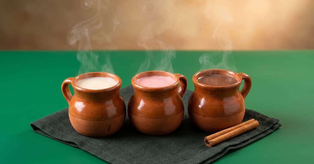 Tres jarritos de barro con atole de vainilla, fresa y chocolate, humeantes, sobre fondo verde.