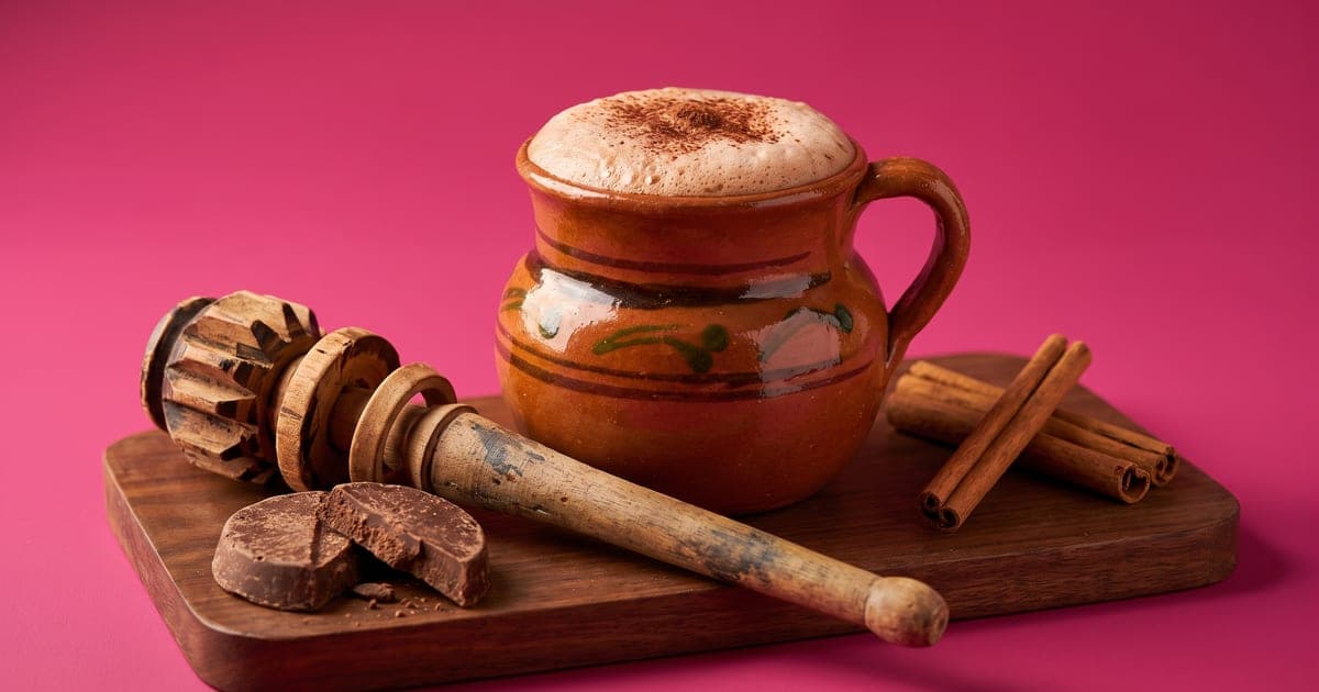 Taza de barro con chocolate caliente espumoso y un molinillo de madera, sobre fondo rosa mexicano.