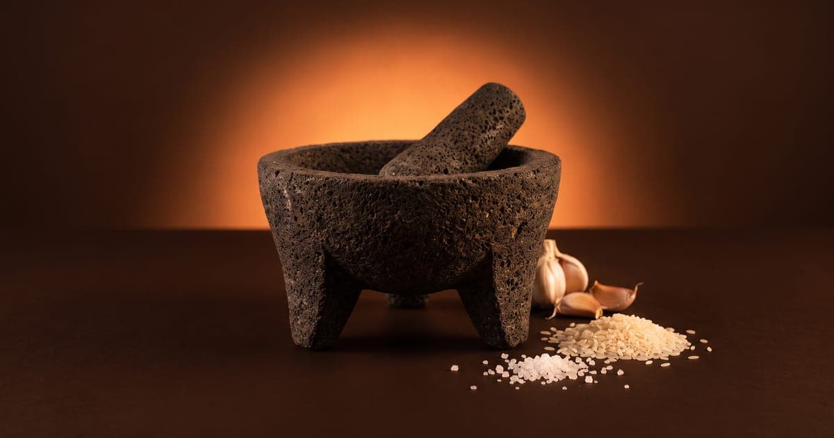 Molcajete de piedra volcánica con su tejolote, dientes de ajo, arroz crudo y sal gruesa, sobre fondo café oscuro.