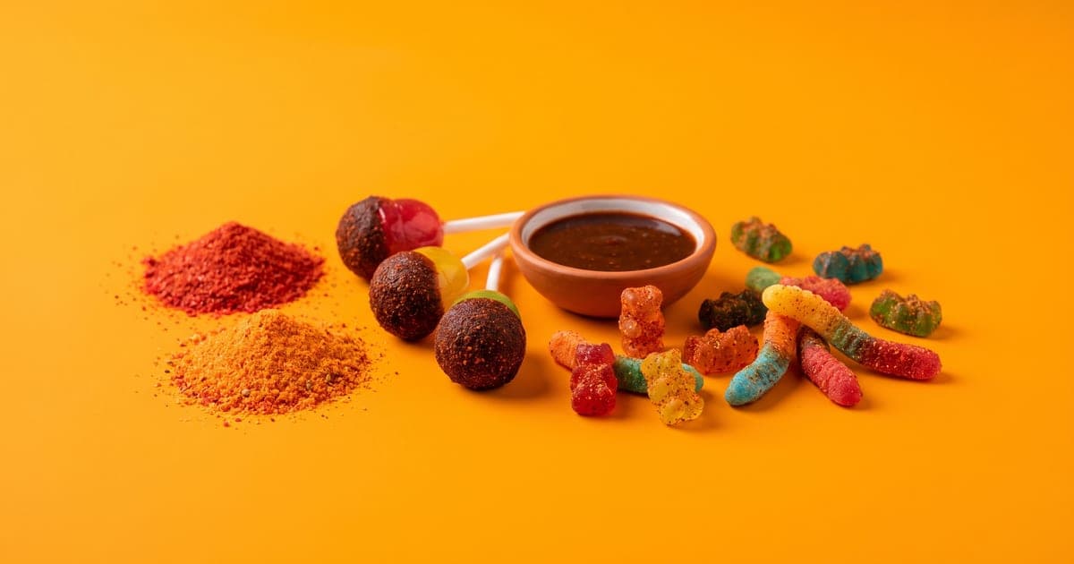 Montoncitos de polvo de dulce enchilado rojo y naranja, paletas de fruta cubiertas de chile, un platito con chamoy oscuro y gomitas enchiladas sobre fondo naranja mango.