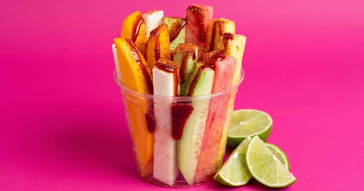 Vaso transparente desbordando bastones de mango, jícama, pepino, sandía y piña bañados en chamoy rojo oscuro y espolvoreados con chile en polvo, con gajos de limón al lado, sobre fondo rosa mexicano liso.