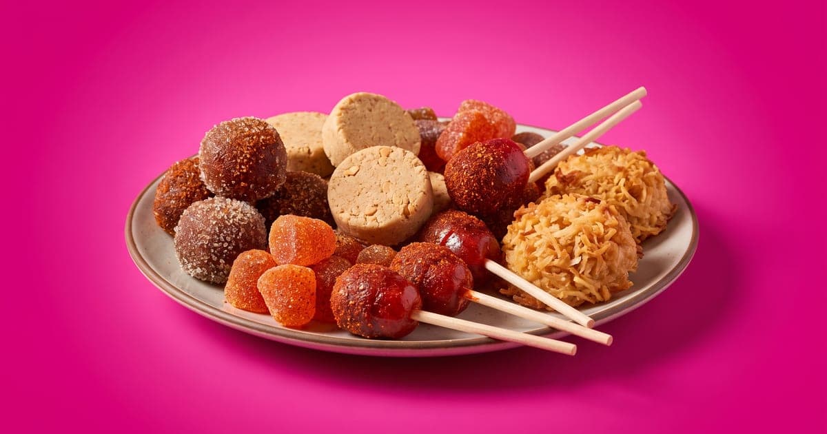 Plato con dulces mexicanos sin envoltura: bolitas de tamarindo con azúcar y chile, mazapanes, paletas enchiladas, gomitas con chile y cocadas, sobre fondo rosa mexicano.