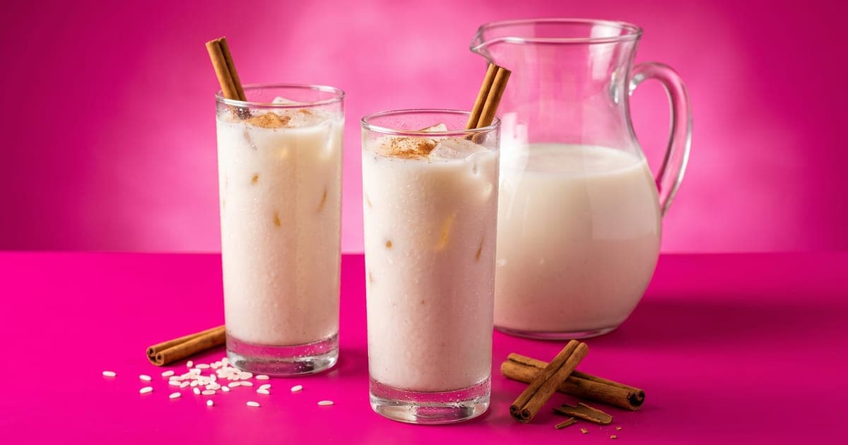 Dos vasos altos de horchata cremosa con hielo y canela molida encima, con rajas de canela dentro, una jarra de vidrio con horchata atrás y granos de arroz y canela al frente, sobre fondo rosa mexicano liso.