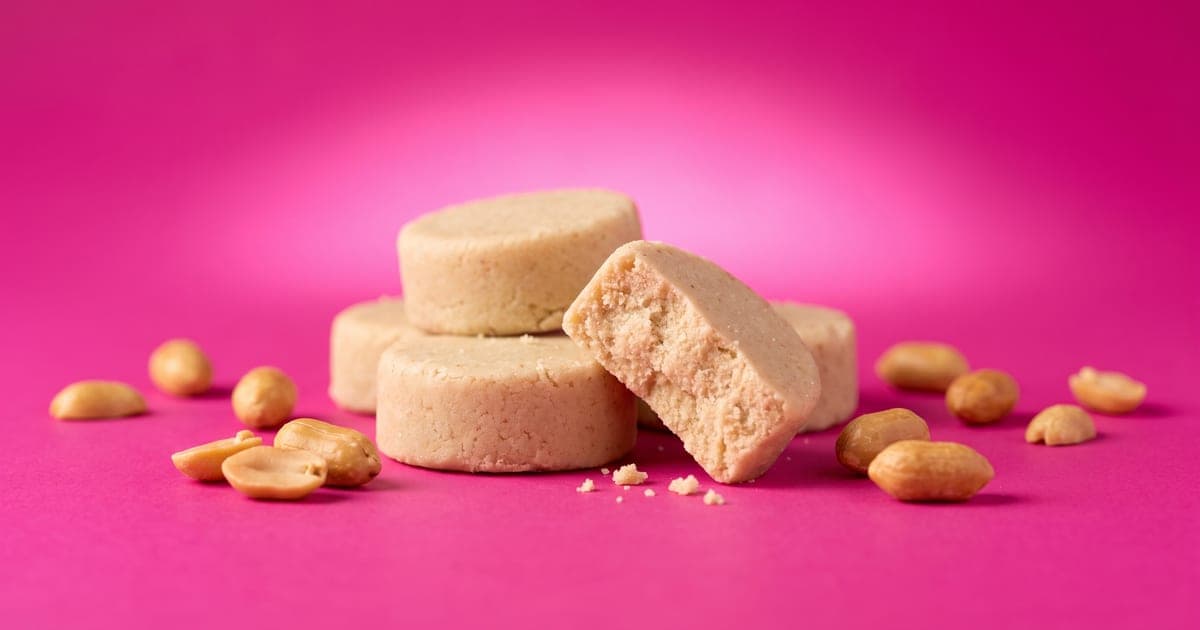 Mazapanes redondos color beige sin envoltura, apilados, uno partido a la mitad mostrando su textura de polvo, con cacahuates tostados alrededor sobre fondo rosa mexicano.