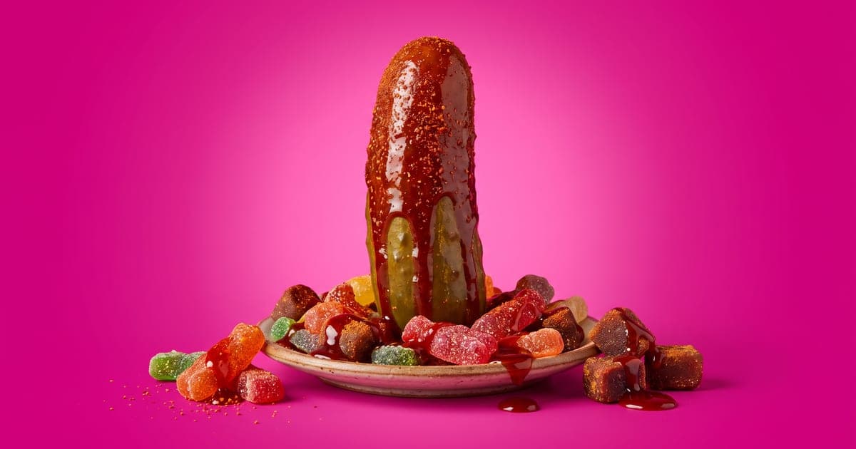 Pepinillo grande bañado en chamoy y chile en polvo con gomitas enchiladas, sobre fondo rosa.