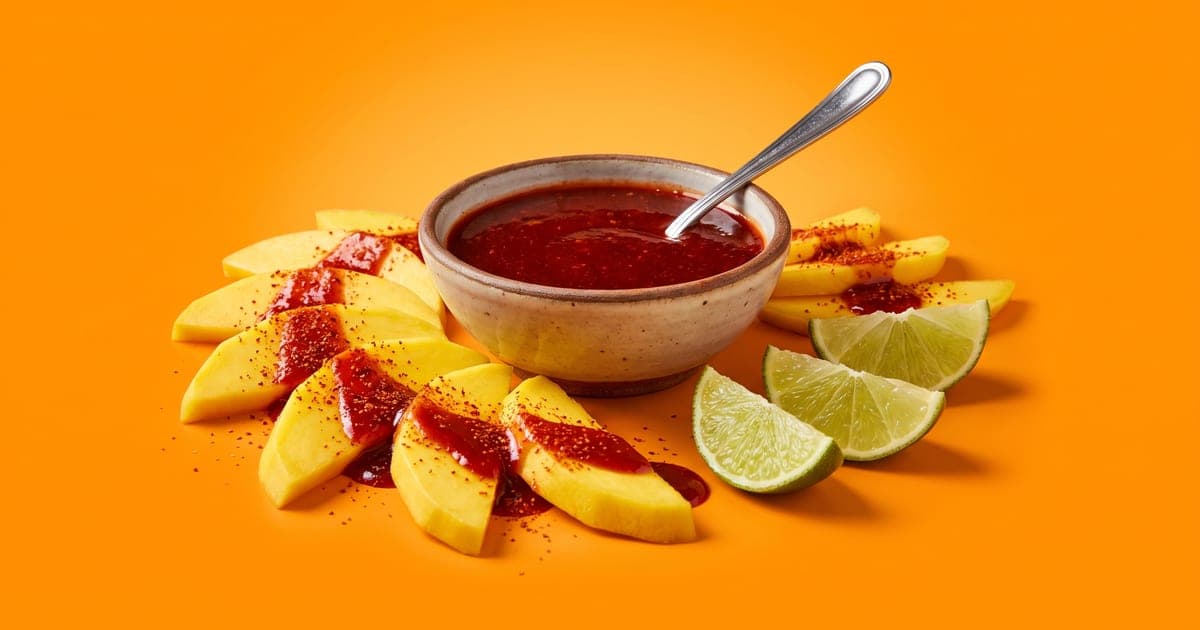 Tazón de chamoy rojo con una cuchara, junto a rebanadas de mango con chamoy y chile en polvo y gajos de limón, sobre fondo naranja.