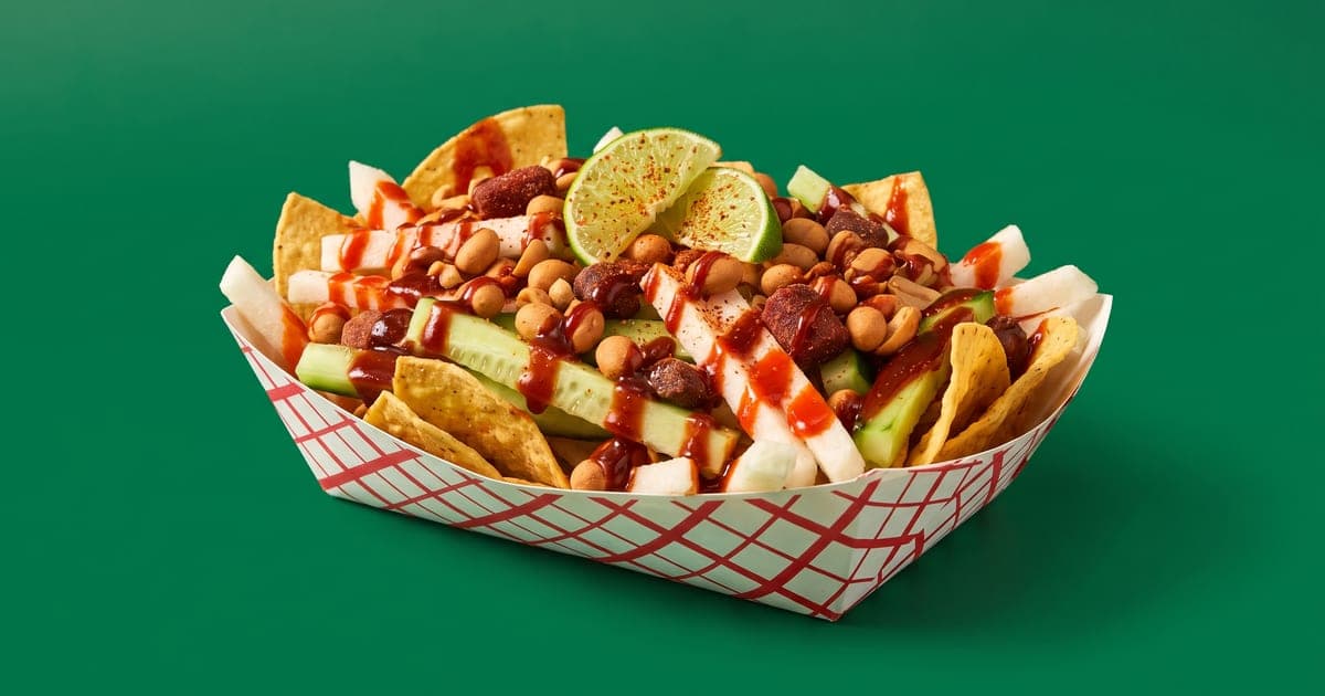 Tostilocos servidos en charola de cartón con pepino, jícama, cacahuates japoneses, chamoy y salsa roja, sobre fondo verde.